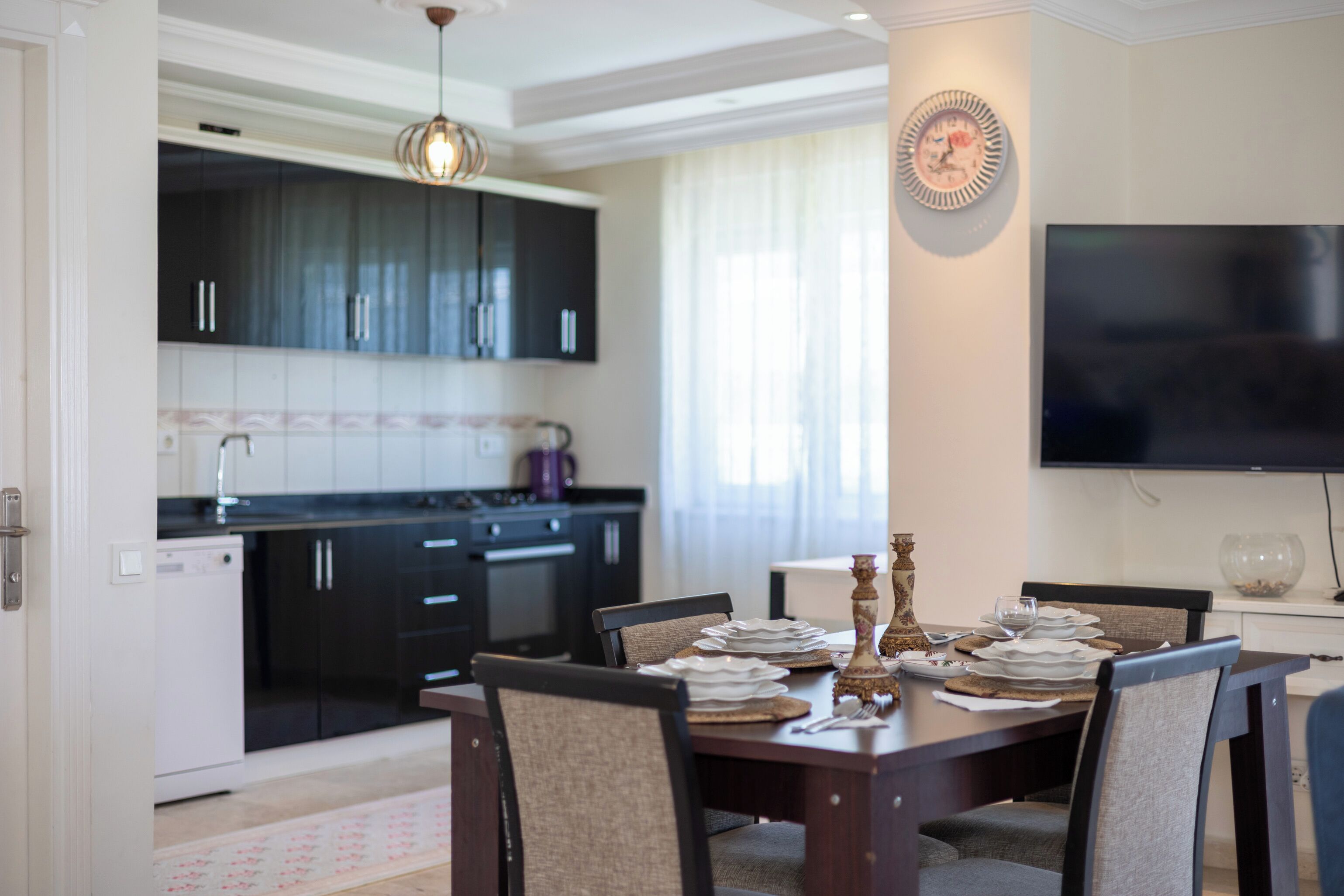 A modern kitchen with dark cabinets, a dining table set for four, and a flat-screen TV mounted on the wall.