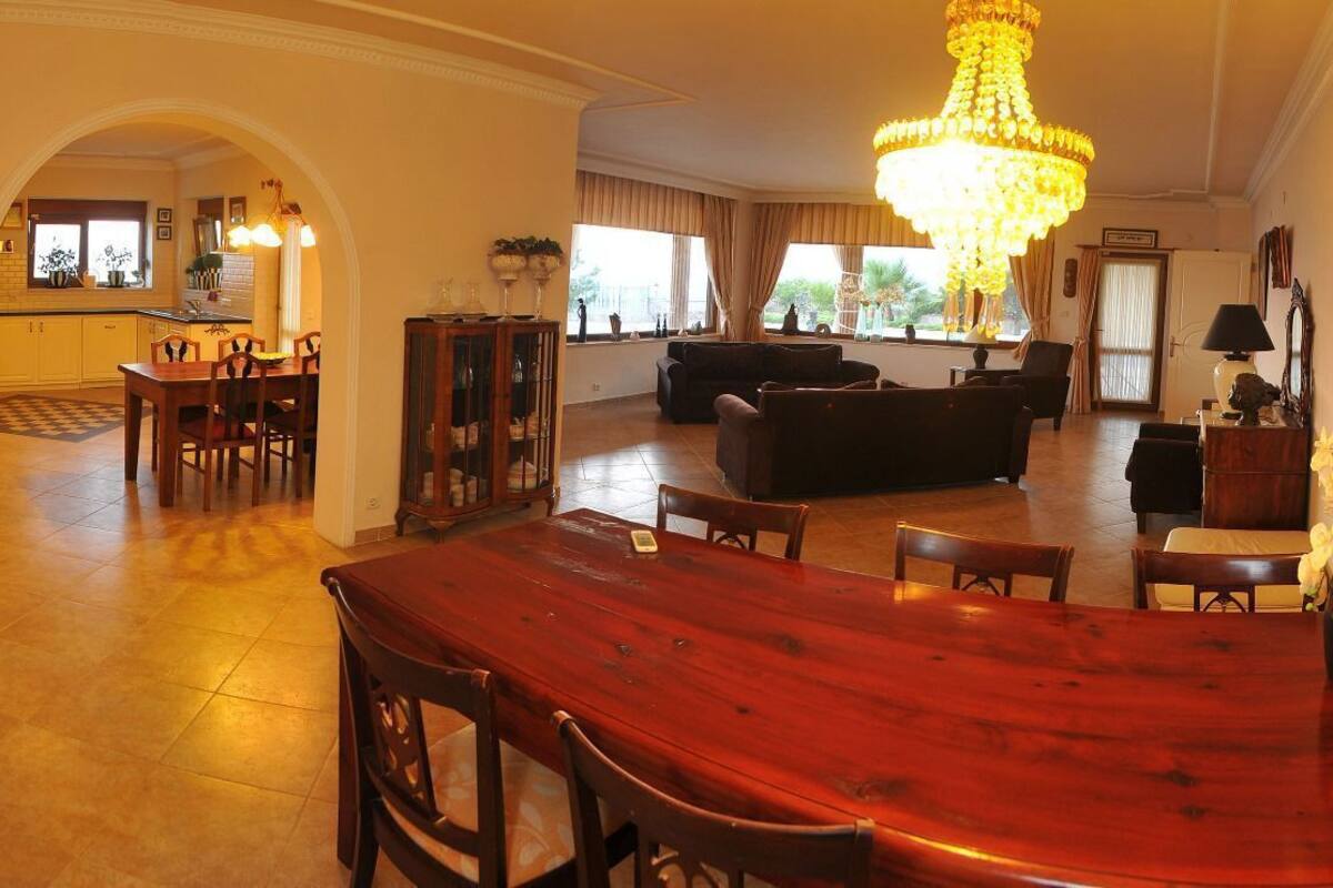 A spacious living area with a wooden dining table, a chandelier, and a view of the kitchen through an archway.