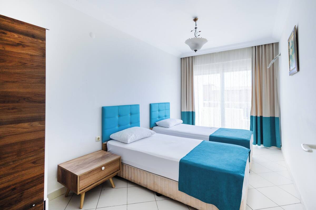 A hotel room with two beds, a wooden wardrobe, a bedside table, and a window with curtains.