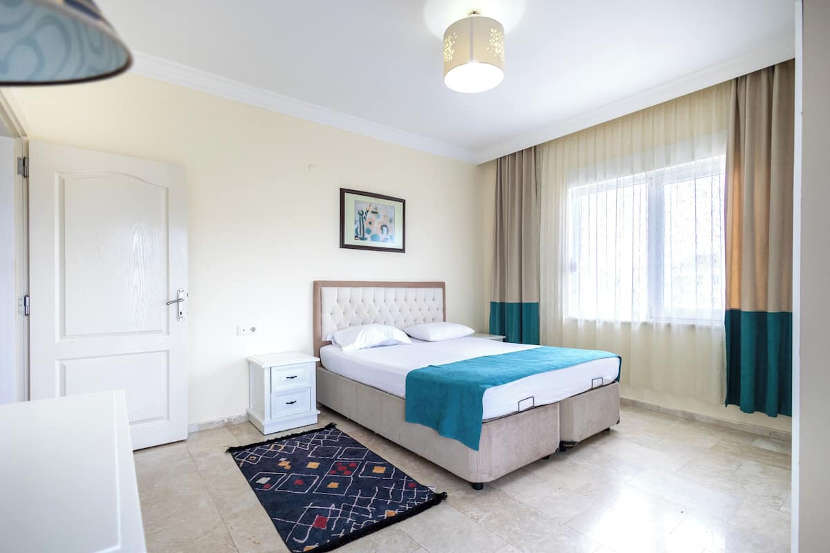 A bedroom with a bed, bedside tables, a nightstand, a window with curtains, and a ceiling light fixture.