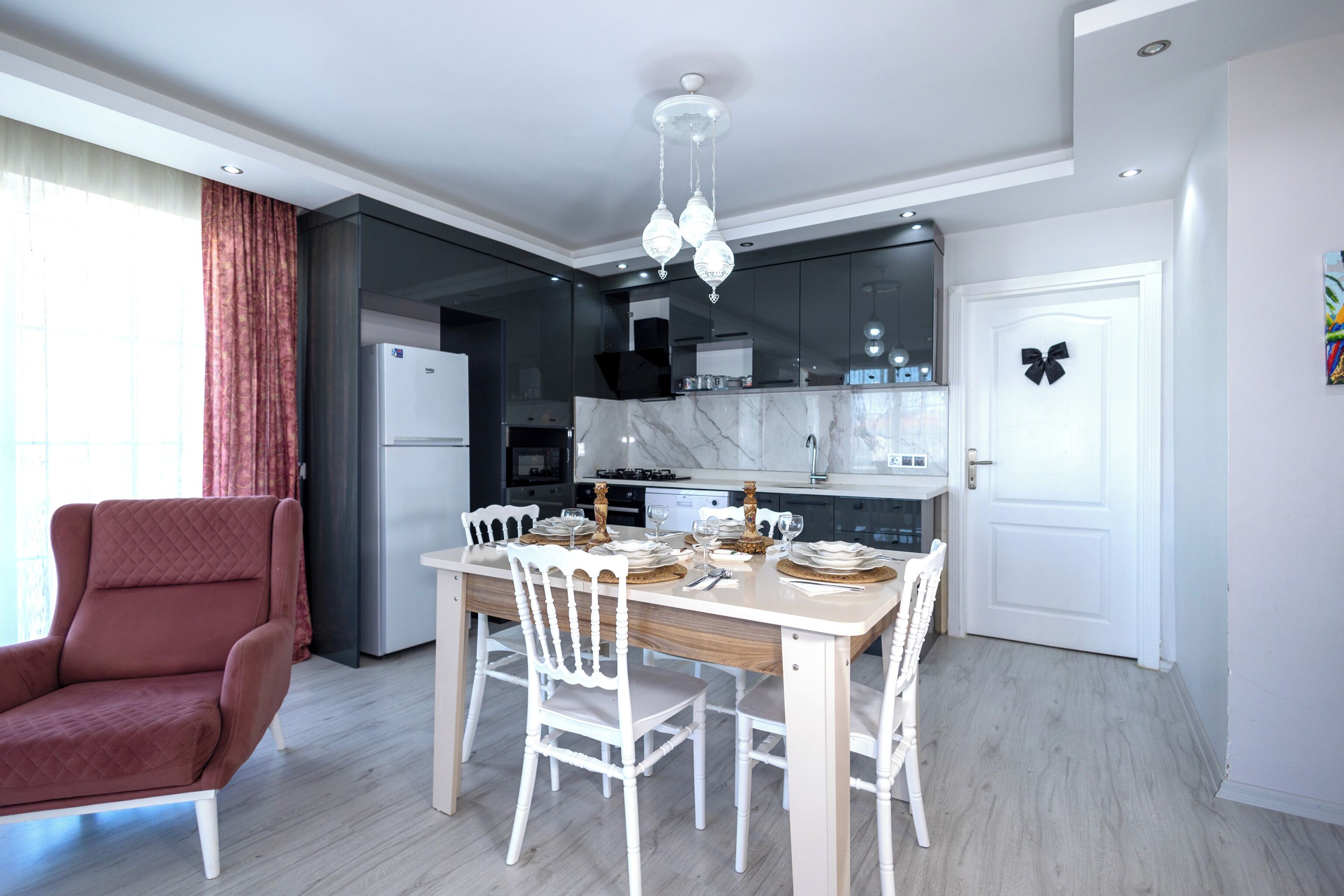 Una cocina moderna con una mesa de comedor preparada para cuatro personas, un sillón rojo y una puerta blanca decorada con un lazo.