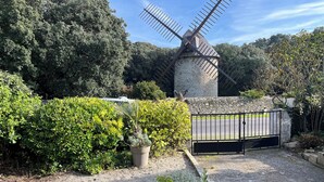 Un moulin à vent en pierre, avec un portail en métal devant, entouré d’une végétation luxuriante et bordé d’un chemin de gravier.