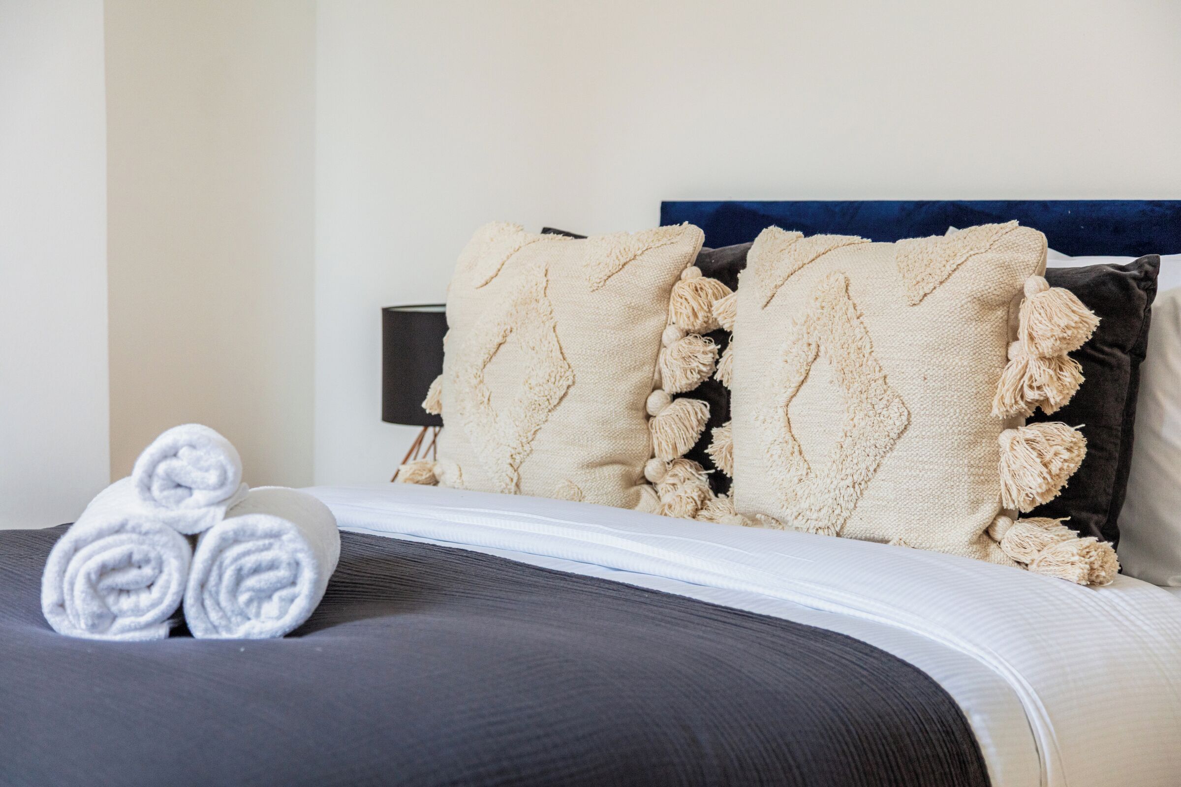 A neatly made bed with rolled towels, a dark headboard, and a wall-mounted lamp.