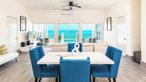 Un vaste espace dédié à la salle à manger, avec une table blanche et des chaises recouvertes d’un tissu bleu, donnant sur un balcon d’où l’on peut admirer l’océan.