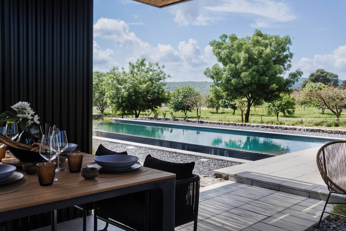 Un área de comedor al aire libre con piscina, mesa, sillas y vistas a árboles y colinas.