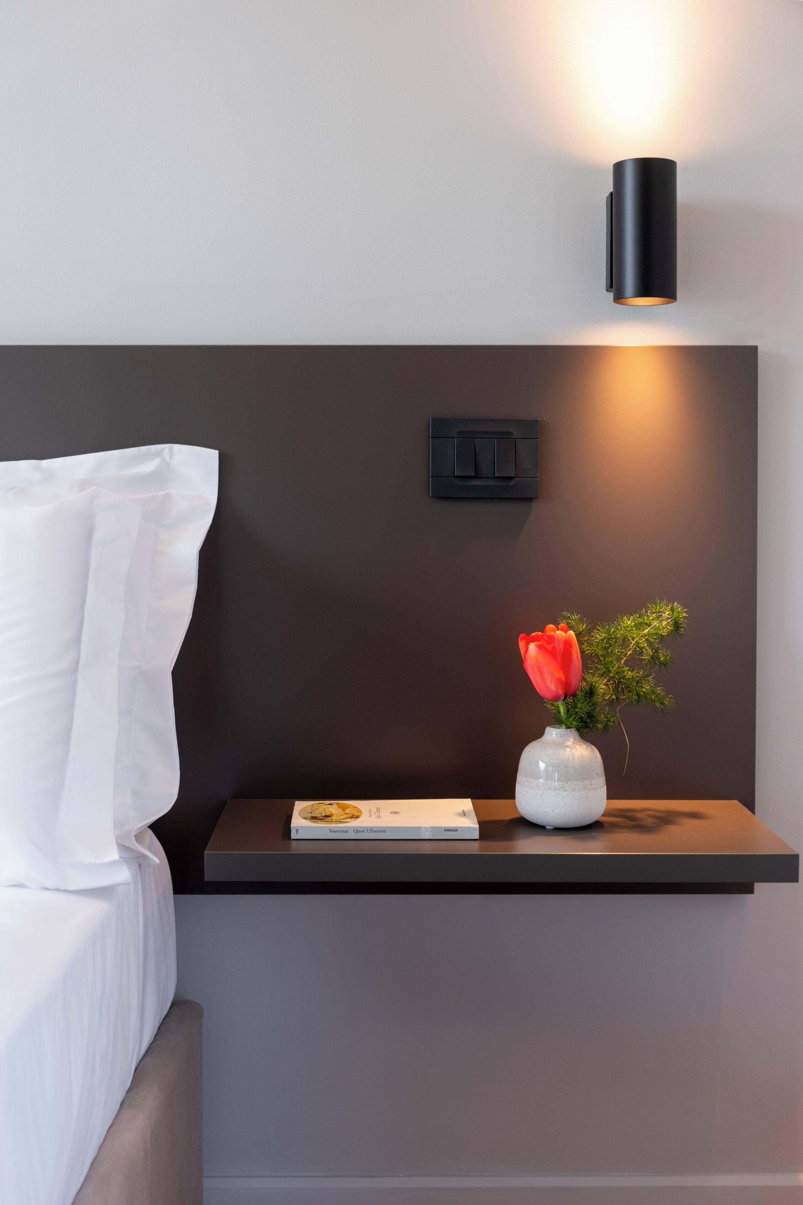 Een moderne hotelkamer met een nachtkastje, een vaas met een rode bloem, een boek en een wandlamp.