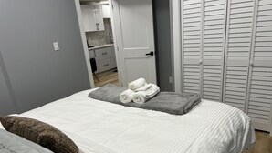 Ein ordentlich bezogenes Bett mit weißer Decke, grauen Decken und einer grauen Tagesdecke, sowie aufgerollten weißen Handtüchern auf dem Bett.