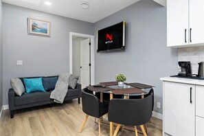 Ein modernes Wohnzimmer mit grauem Sofa, schwarzen Sesseln, einem Glastisch, einem an der Wand befestigten Fernseher und einer Küchenzeile mit Schränken.