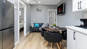 Eine moderne Küche mit Einbauofen, ein Essbereich mit Tisch und Stühlen, ein graues Sofa mit Kissen sowie ein an der Wand befestigter Fernseher.