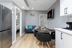 A modern kitchen with a built-in oven, a dining area with a table and chairs, a grey sofa with cushions, and a TV mounted on the wall.