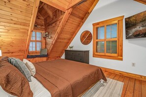 Een slaapkamer met een schuine plafond, een bed met een bruine sprei, een dressoir, een raam met luiken, en een schilderij aan de muur.