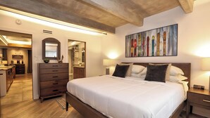A bedroom with a large bed, wooden headboard, bedside lamps, a painting on the wall, and a view into the kitchen.