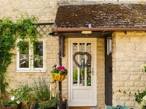 A stone house with a white door, a heart-shaped wreath, and various potted plants. - Cosy Cotswolds home | parking | colourful garden | Pass the Keys (Bourton-on-the-Water)