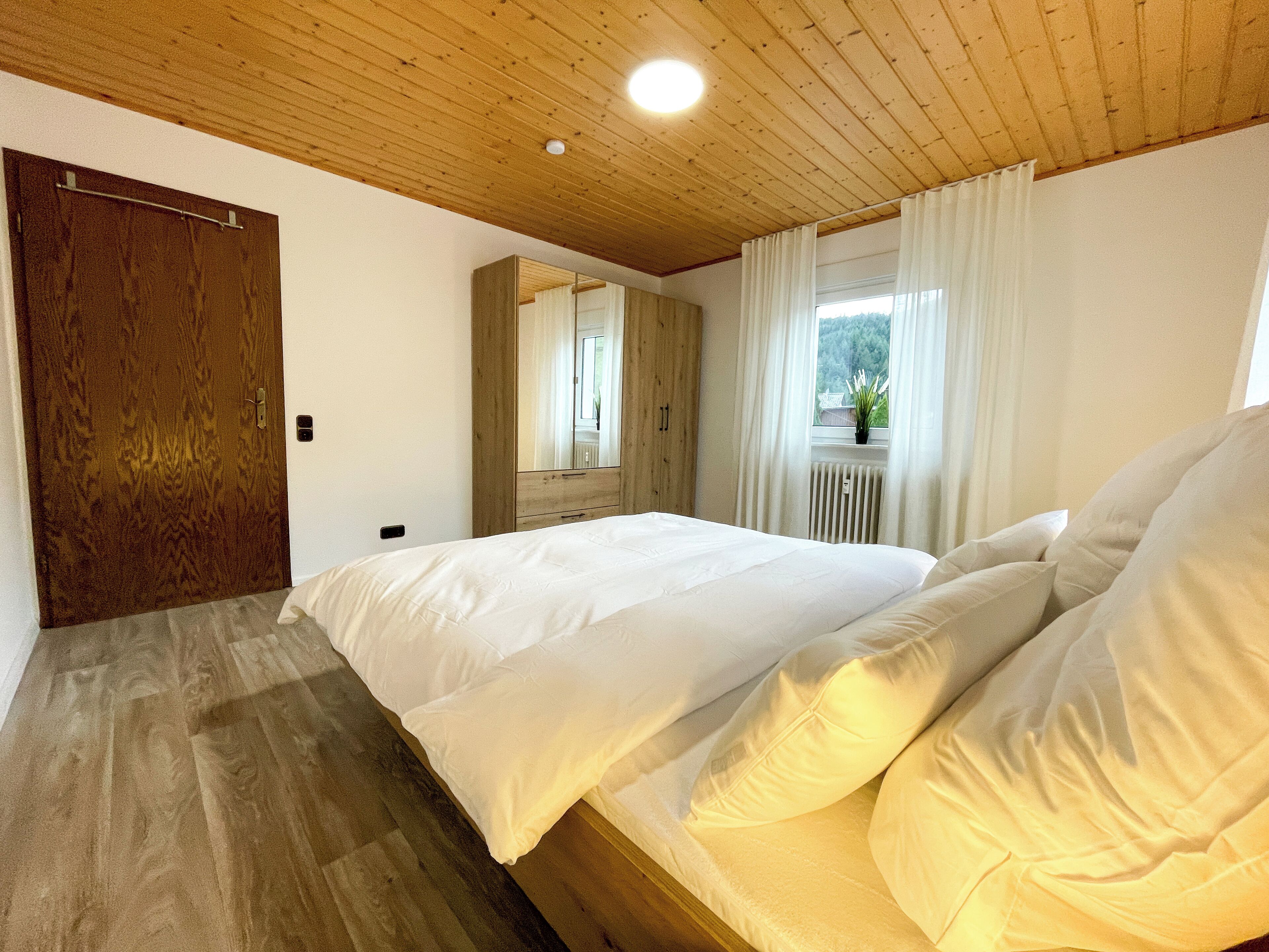 A bedroom with a wooden ceiling, a large bed with white linens, a wooden wardrobe, and a window with curtains.