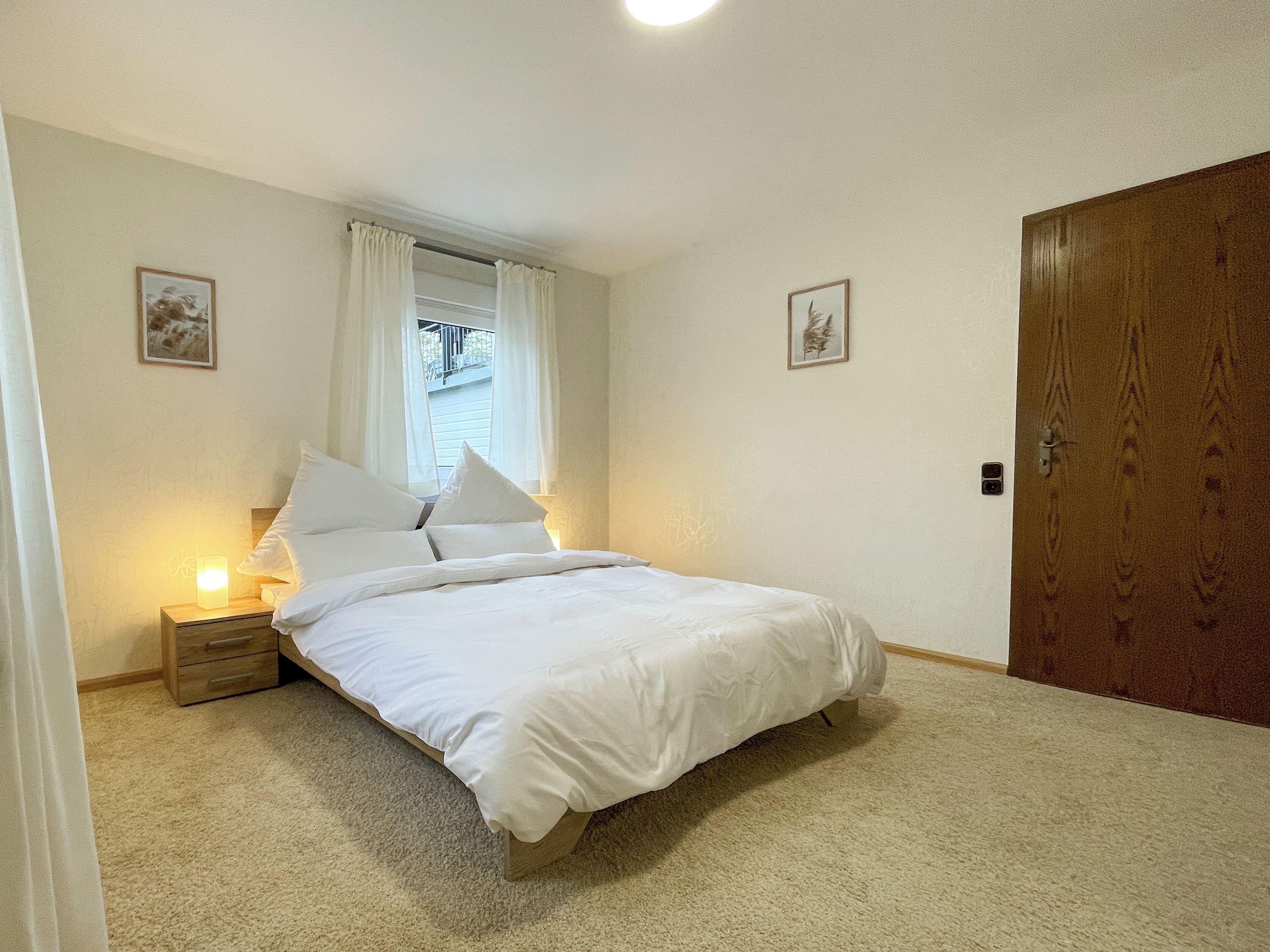 Quarto com uma cama, uma mesinha de cabeceira, uma janela com cortinas, uma porta e duas molduras com quadros na parede.