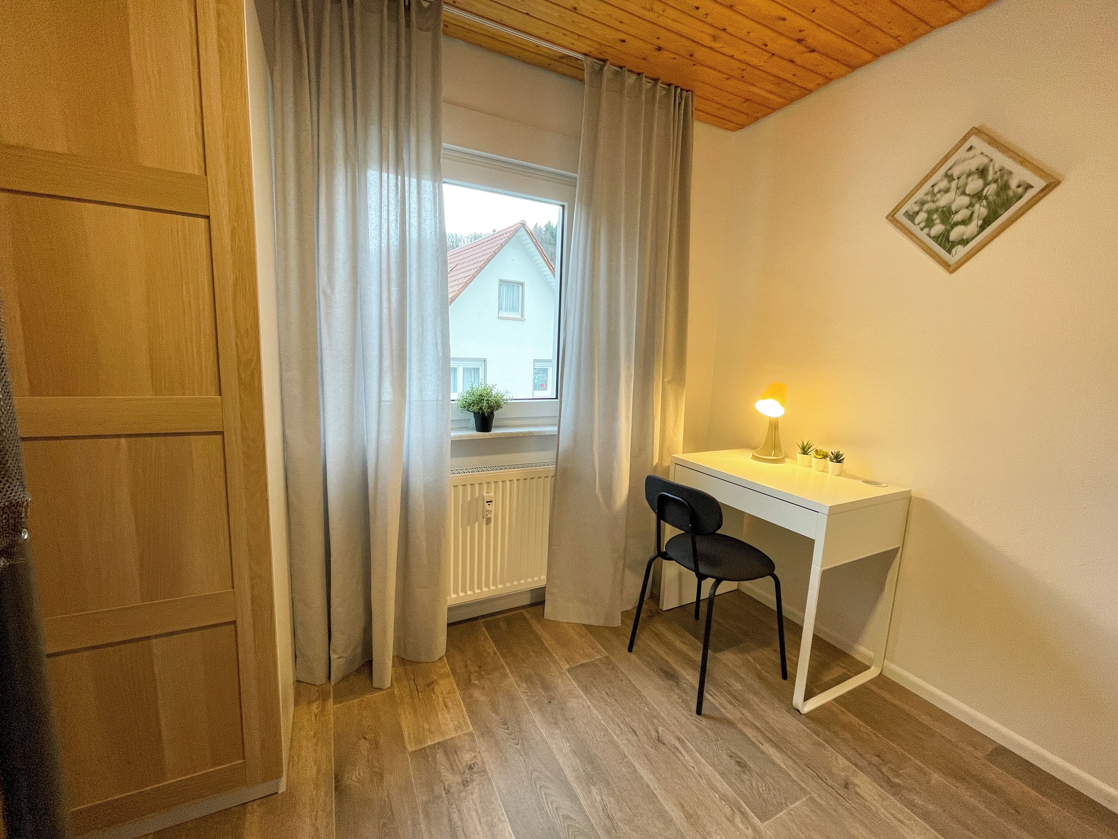 Une pièce avec un plafond en bois, un bureau blanc avec une chaise, un radiateur et une fenêtre avec des rideaux.