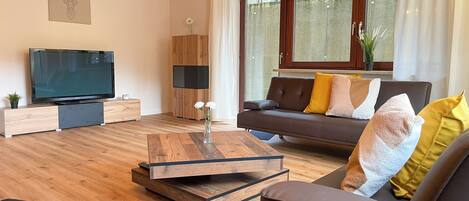 Ein modernes Wohnzimmer mit Flachbildfernseher, Holzboden, einem braunen Sofa mit Kissen, einem Couchtisch und einem großen Fenster mit Vorhängen.