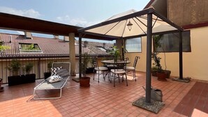 Een overdekt terras met een tafel, stoelen, een loungestoel en potplanten.
