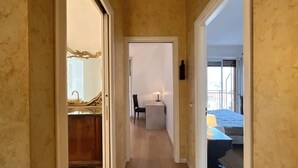 Een smalle gang die leidt naar een kamer met een bed, een bureau en een stoel.