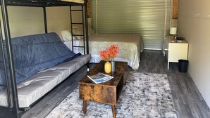 Una habitación compacta con una cama litera, un sofá, una pequeña mesita de noche con un jarrón de flores y una persiana cerrada.