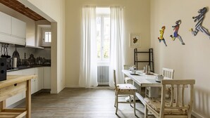 Une cuisine avec des meubles blancs, une table en bois et des chaises. Il y a une fenêtre avec des rideaux, un radiateur et un tableau accroché au mur.