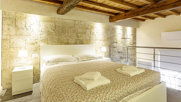 Une chambre avec un mur en pierre, des poutres en bois au plafond, un lit recouvert d’une couvre-lit à motifs floraux et une table de chevet avec une lampe.
