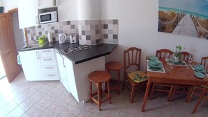 A compact kitchen with a microwave, stove, and sink. Adjacent is a dining area with a wooden table set for four, chairs, and a small potted plant.