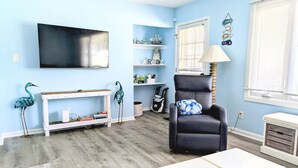 A living room with a flat-screen TV, a black leather chair, a white console table, and a bookshelf with decorative items. - Cozy White Cottage by the Bay (Lower Township)