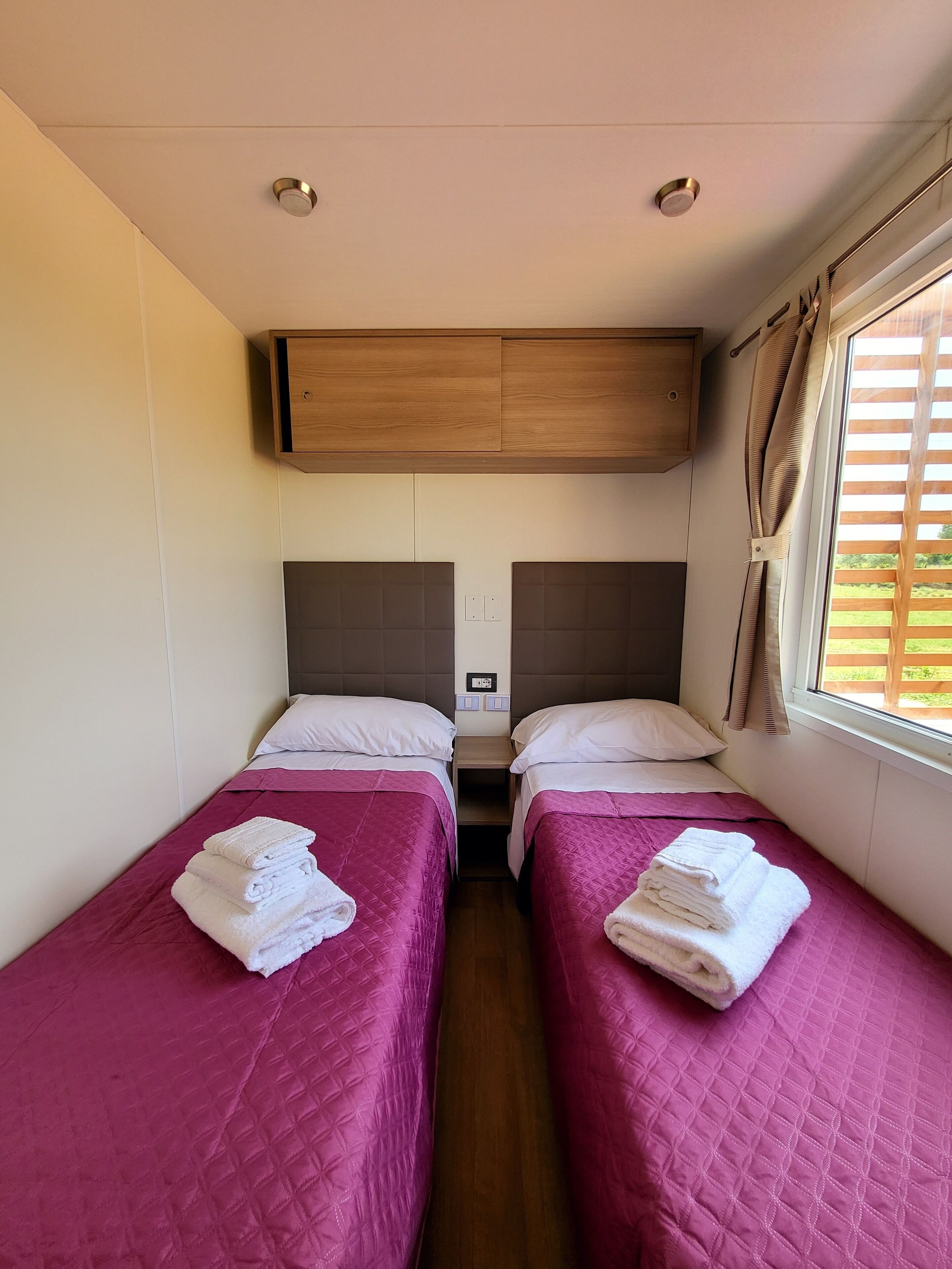 Une chambre avec deux lits, chacun recouvert d’une courtepointe rose matelassée et agrémenté d’oreillers blancs, et dotée d’une tête de lit en bois. Chaque lit est muni de serviettes, et une fenêtre avec des rideaux se trouve sur le côté droit.