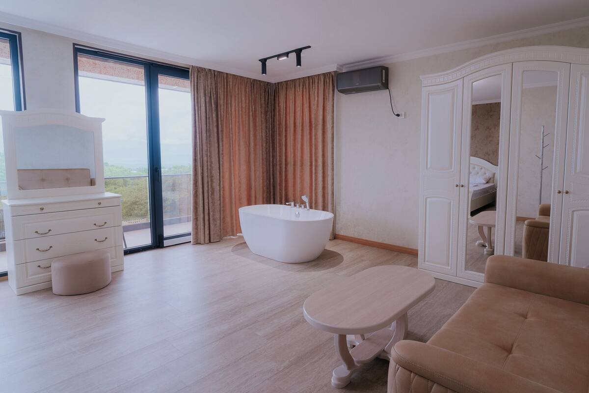 A modern hotel room with a large window, a white bathtub, a beige sofa, and a wardrobe with a mirror.