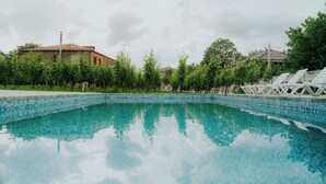 A swimming pool with clear blue water, surrounded by trees and a building with a tiled roof. - Villa Ikalto (Telavi)