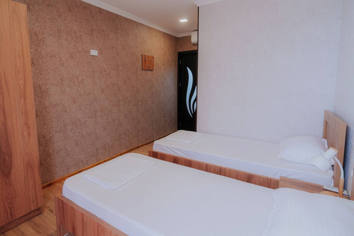 A room with two single beds, wooden wardrobe, textured wall, and a small wooden panel on the wall.