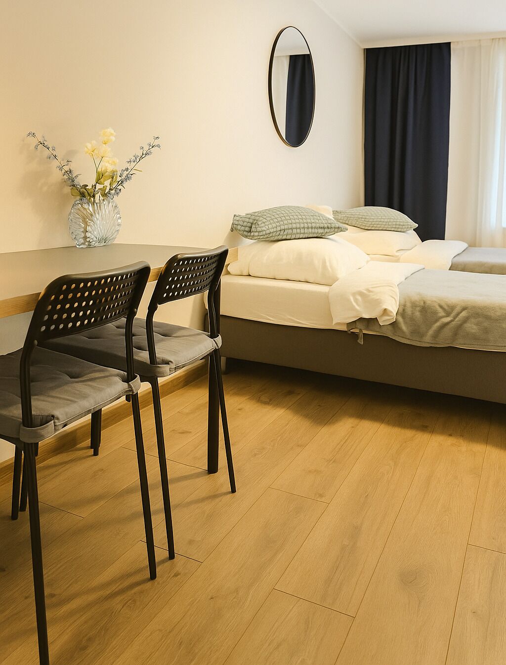 Ein Zimmer mit einem Bett, zwei Stühlen, einem kleinen Tisch, einer Vase mit Blumen und einem Spiegel an der Wand.