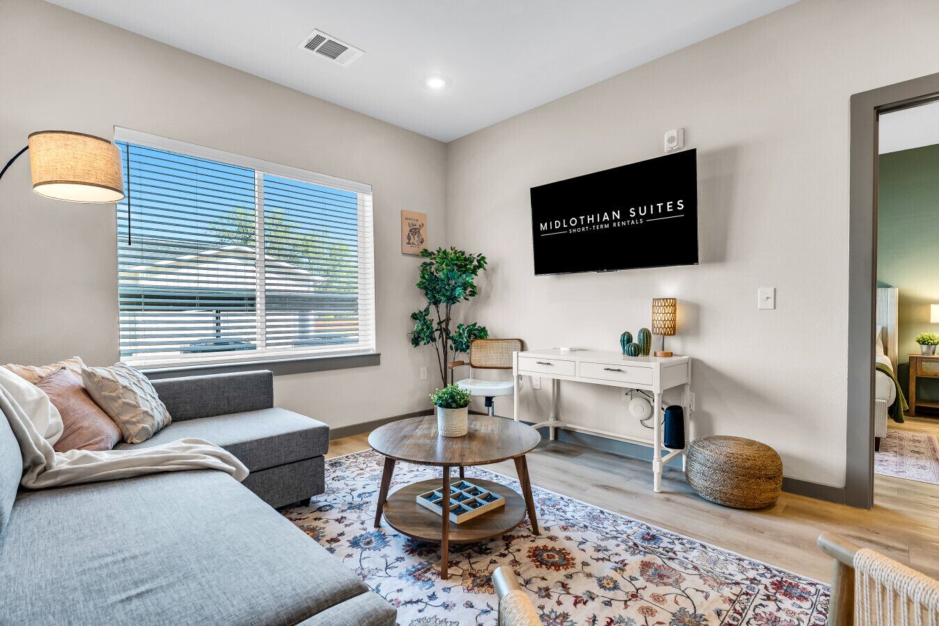 A modern living room with a grey sofa, a round coffee table, a white desk, and a flat-screen TV mounted on the wall.