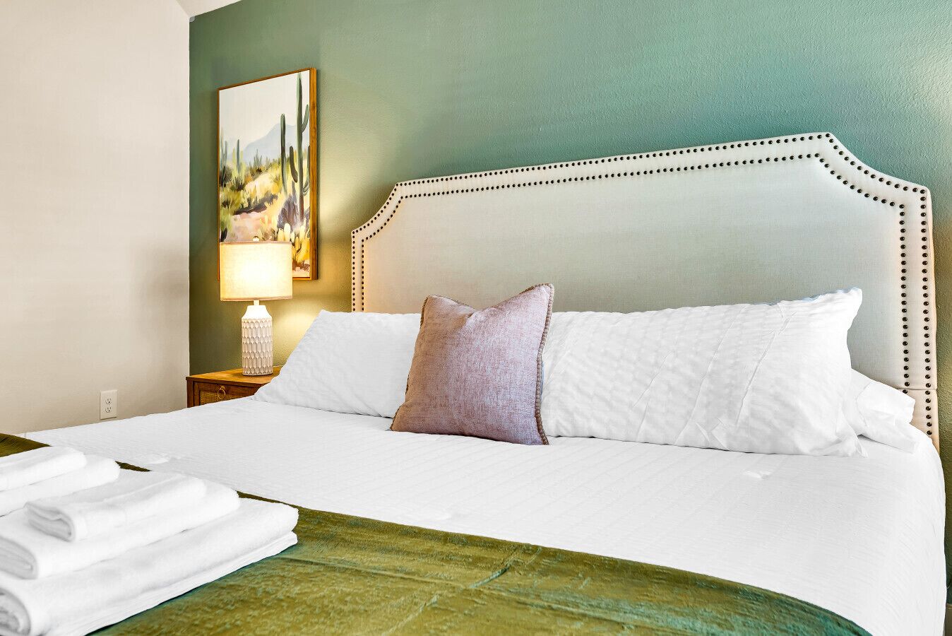 Una cama bien hecha con sábanas blancas y una sola almohada decorativa, una lámpara de noche y una toalla doblada en el borde de la cama.