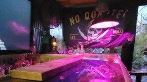 A bar area with a wooden floor, a neon skull sign, and a large screen displaying "NO QUARTERS".