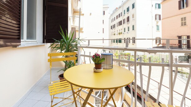 A balcony with a yellow table and chairs, potted plants, and a view of adjacent buildings.