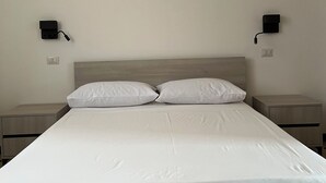 Un lit bien fait, avec du linge de lit blanc, deux tables de chevet et des lampes de chevet fixées au mur.