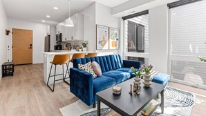 Un salon moderne avec un canapé bleu, une table basse et un coin cuisine doté de meubles blancs et de chaises orange.