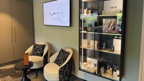 A modern interior with a large display case of luxury brands, two armchairs with patterned cushions, and a wall-mounted screen displaying 'LA CÔRNICHE'. - LaCorniche (Contrexéville)