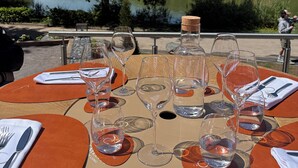 A table set for a meal with wine glasses, water bottles, and cutlery, overlooking a lake with trees and a person in the background. - LaCorniche (Contrexéville)
