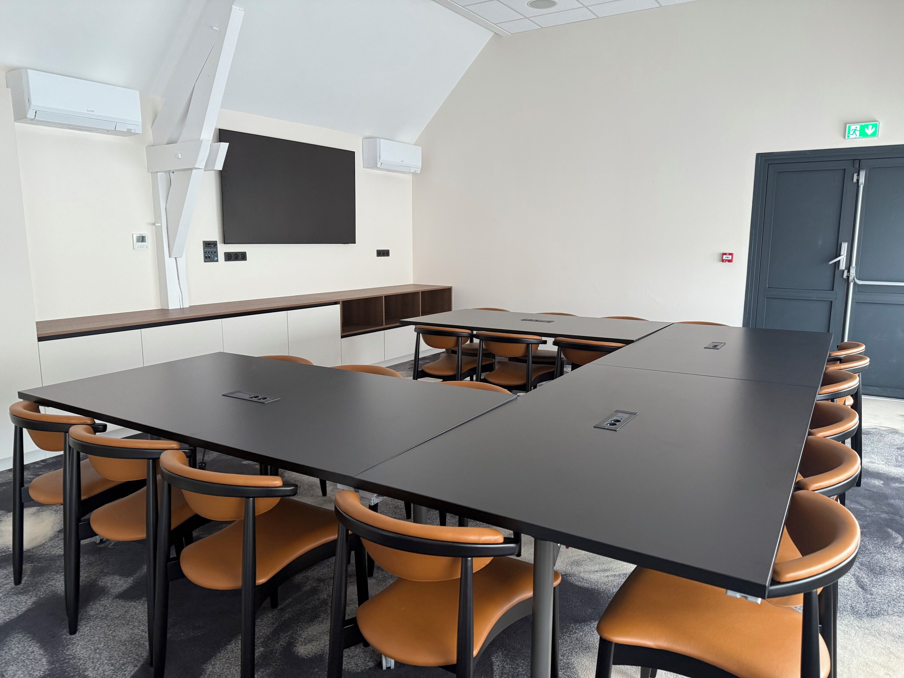 Un salón de conferencias con una gran mesa, varias sillas, una pantalla de proyector y una puerta con letrero de salida de emergencia.