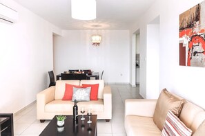 A living room with a sofa, a coffee table with a bottle and glasses, a dining area with chairs and a table, and a kitchen in the background.