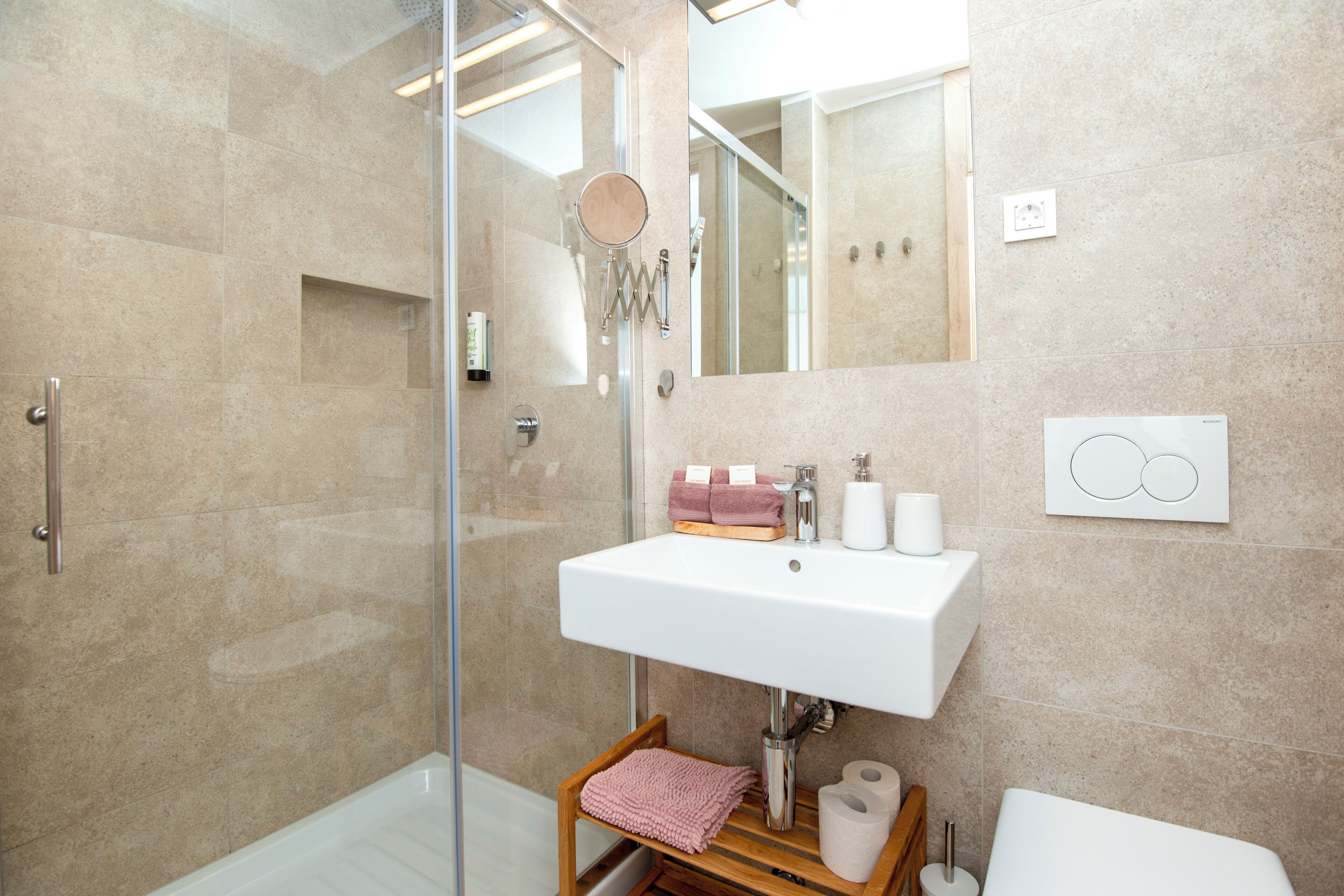 Ein modernes Badezimmer mit einer Duschkabine aus Glas, einem weißen Waschbecken mit Spiegel darüber und einem Holzregal mit Handtüchern.