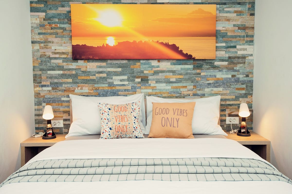A bedroom with a stone mosaic wall, a bed with striped bedding, two bedside lamps, and two pillows with motivational phrases.