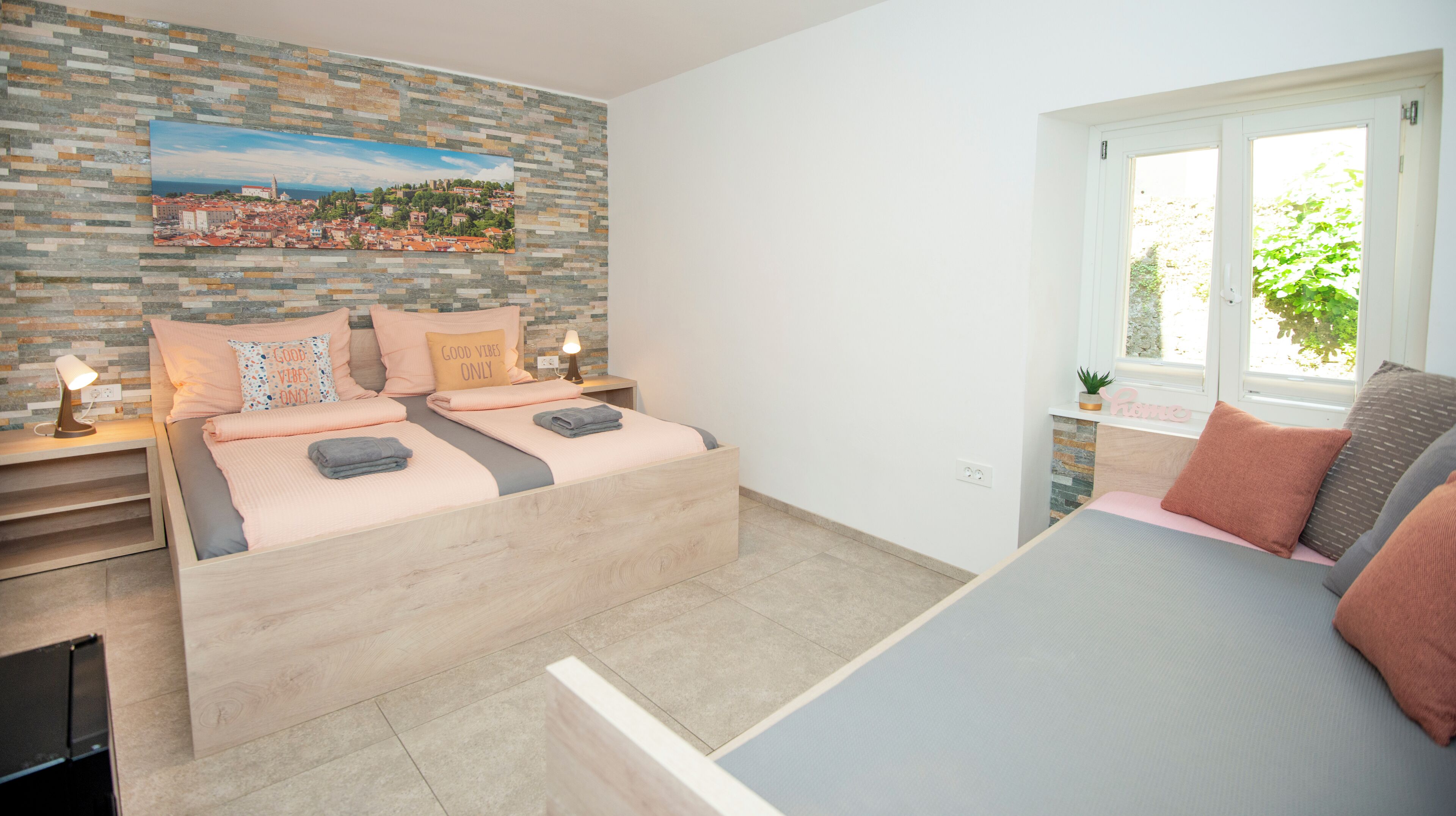 Un dormitorio con dos camas, una pared de piedra con un cuadro de paisaje urbano, una ventana con vistas a la vegetación y una mesita de noche con una lámpara.