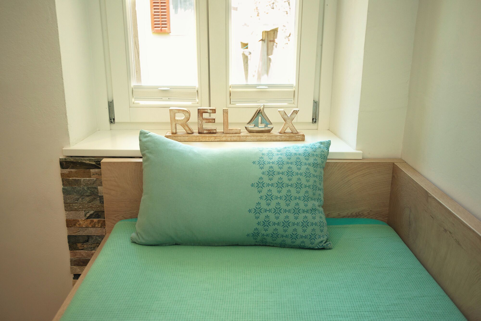 A bed with a green bedspread and a pillow with a blue pattern, a window with shutters, and a wooden shelf with the word "RELAX" and a decorative item.