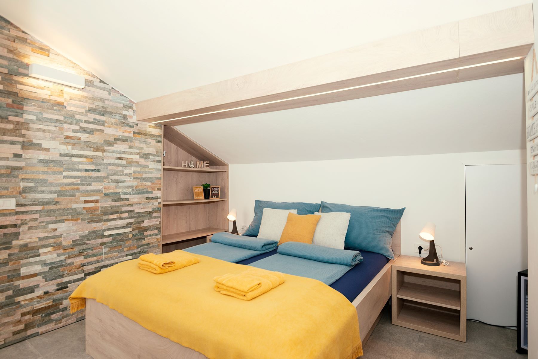 A bedroom with a sloped ceiling, a stone wall, a wooden shelf with a 'HOME' sign, a bed with blue and yellow bedding, and a bedside table with a lamp.