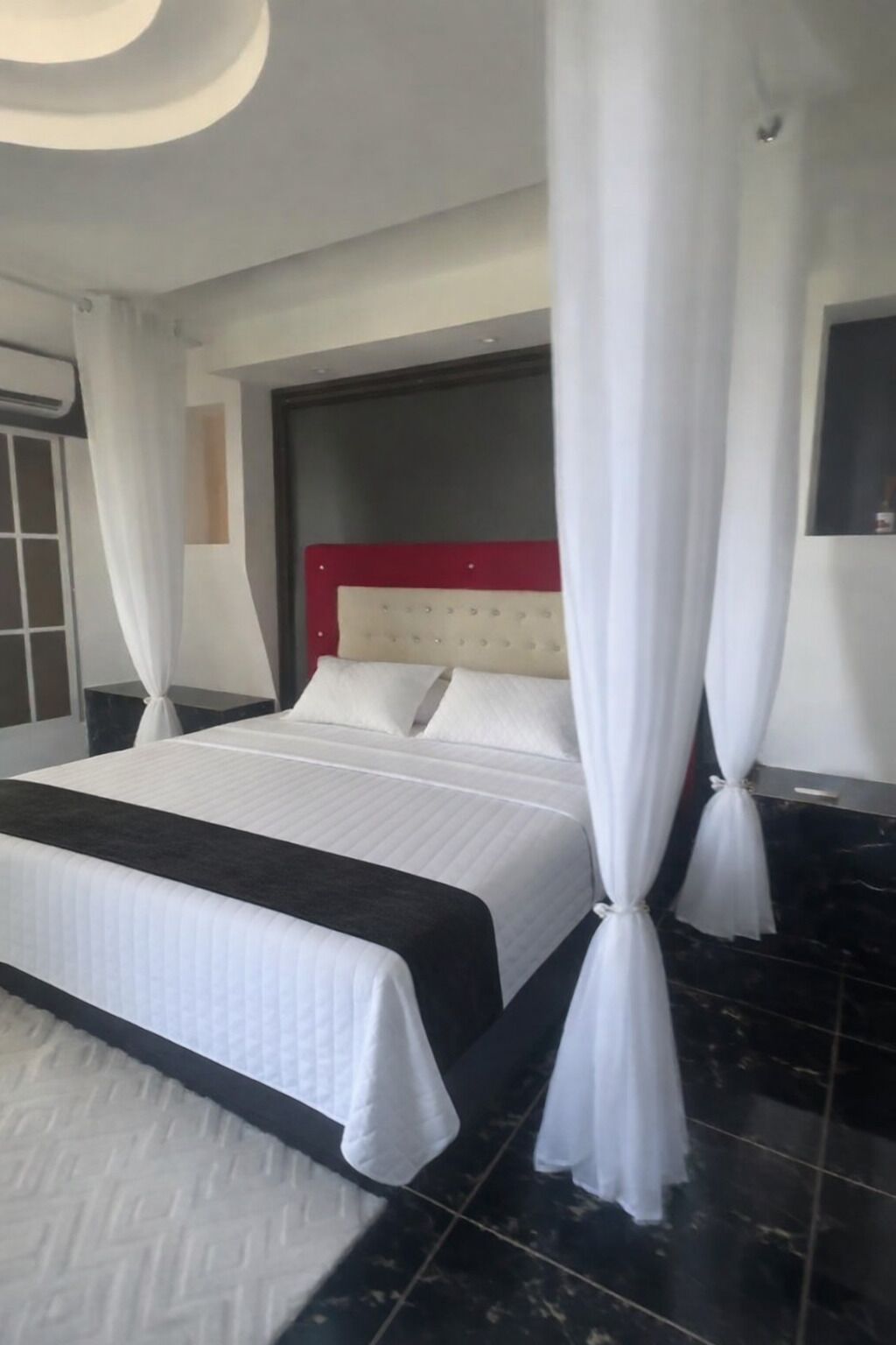 A bed with white bedding, a red headboard, and white curtains.