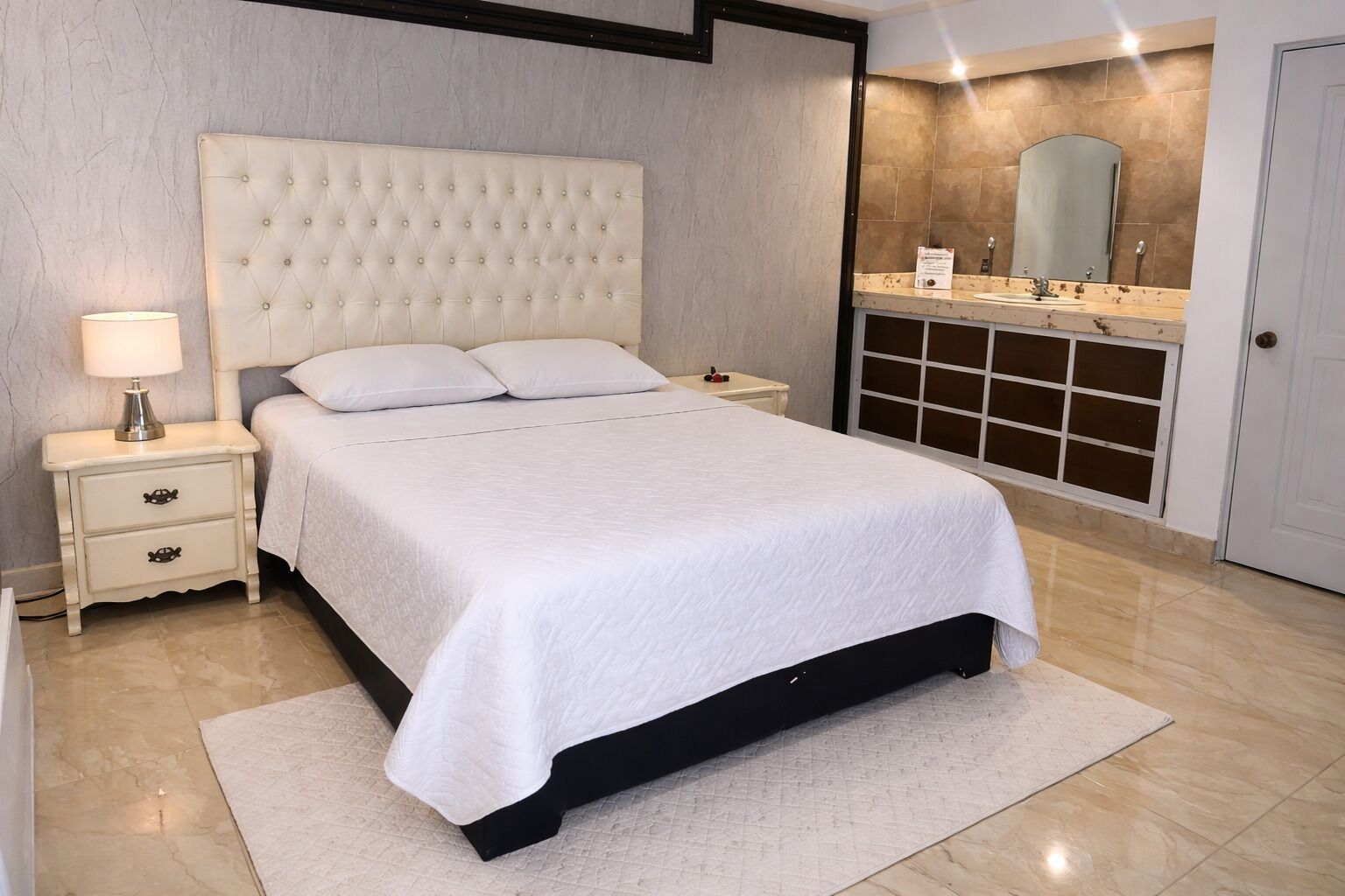 Ein Schlafzimmer mit einer gebügelten Kopfteil, einem Nachttisch mit Schubladen, einem Beistelltisch mit Lampe, einem großen Bett mit weißer Bettwäsche, einem Badezimmer mit Spiegel und Waschbecken sowie einer Tür.